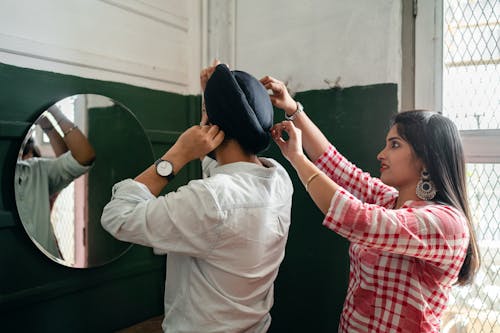 Side view of positive wife wearing plaid dress and earrings in authentic design adjusting turban on head of young Sikh husband in denim shirt standing together against mirror on wall