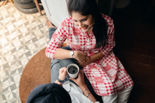 From above of cheerful young wife in checkered tunic with bindi on forehead sitting in doorway against street kitchen leaning on knees of husband in turban while spending free time together at home