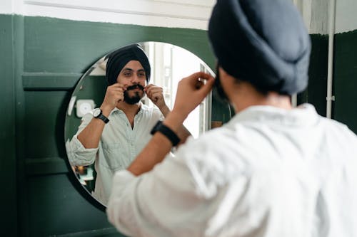 Back view of handsome young Indian male hipster with twisted mustache wearing denim shirt and traditional turban standing against mirror on wall while taking care about appearance