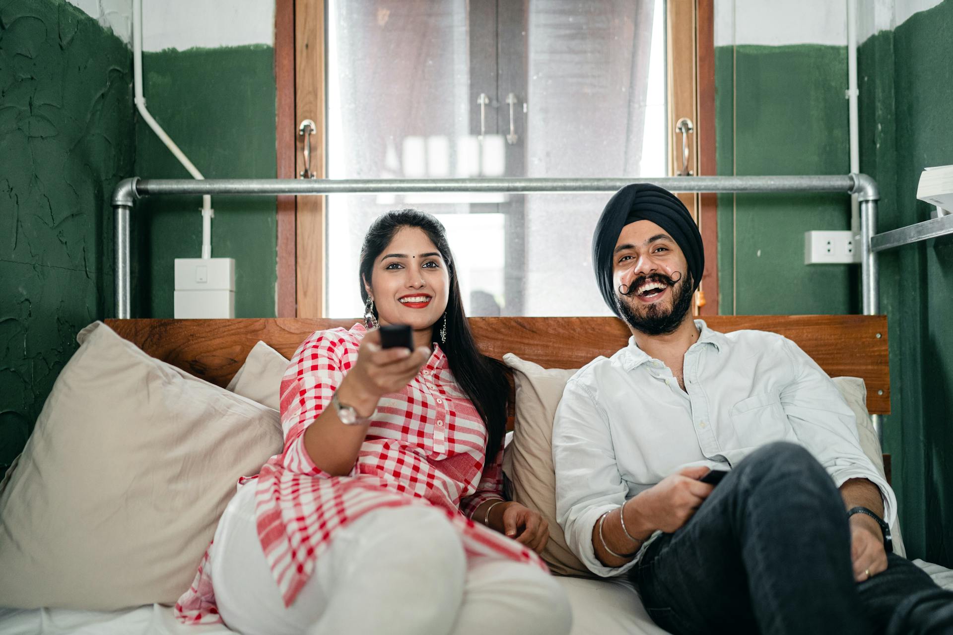 Cheerful wife with bindi on forehead wearing plaid tunic with white trousers using TV remote control for channel switching while lying on bed with laughing Sikh husband in turban with stylish beard and twisted mustache