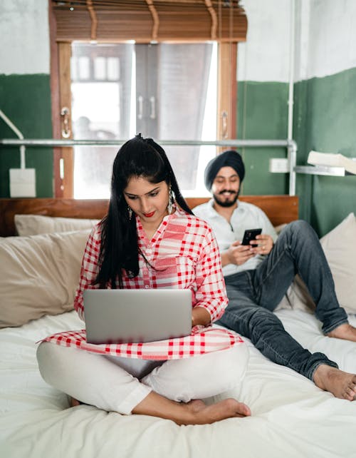 Free Young Indian man in casual clothes and turban resting on bed and browsing mobile phone while young Indian woman sitting in lotus pose and typing on netbook in cozy bedroom Stock Photo