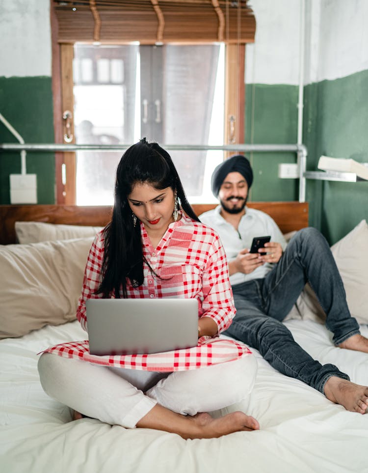 Ethnic Couple Using Laptop And Smartphone On Bed At Home