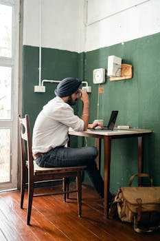 Side view of concentrated Indian guy with dark beard in turban using laptop while working remotely in contemporary apartment at daytime