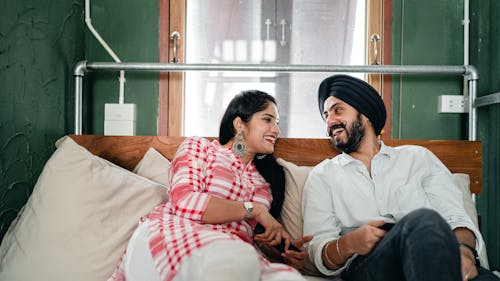 Cheerful young Indian man and woman laughing and looking at each other while enjoying pastime on sofa in modern apartment