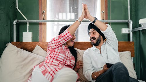 Positive adult ethnic couple in casual clothes giving high five to each other while resting on bed in small bedroom with sticking out pipe