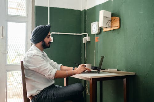 Side view of focused adult ethnic man in casual clothes and hat using netbook while sitting at table with pen