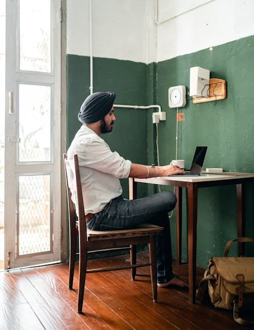 Free Side view of full length concentrated bearded and barefoot ethnic man in casual clothes using netbook while sitting at table at home Stock Photo