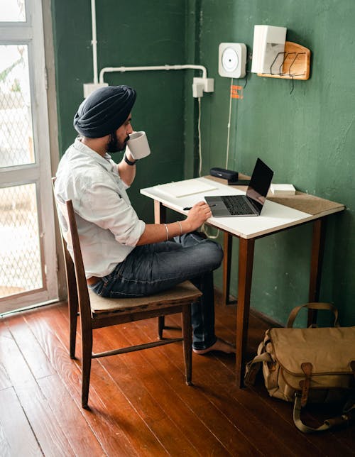 Free From above side view of full length barefoot and bearded focused ethnic male in casual clothes using laptop while sitting at table at home crossing leg and drinking tea and holding pen for writing notes in planner Stock Photo