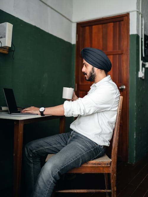 Side view of concentrated bearded ethnic male in casual clothes and hat using netbook at home while sitting at table and drinking cup of hot tea