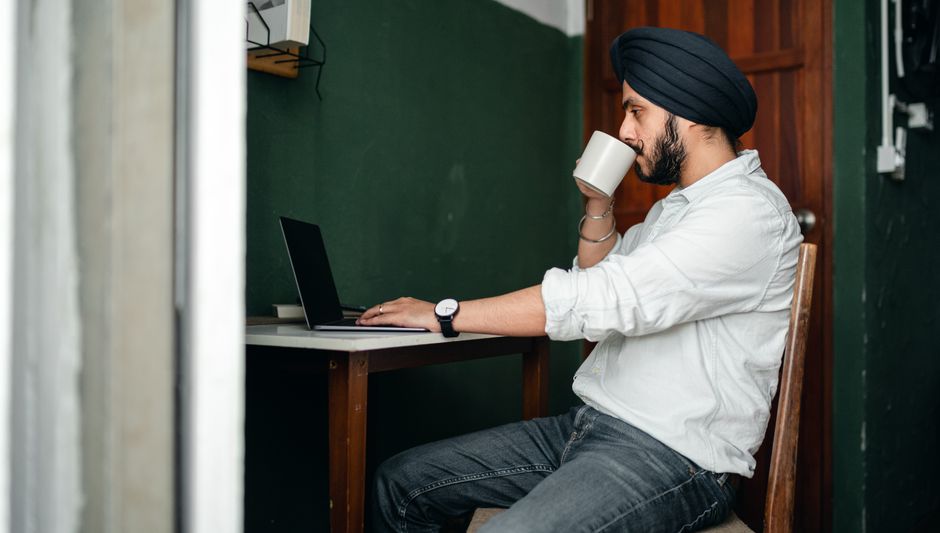 Side view of concentrated bearded male in casual outfit and hat using netbook at home while sitting at table and drinking tea Side view of concentrated bearded male in casual outfit and hat using netbook at home while sitting at table and drinking tea