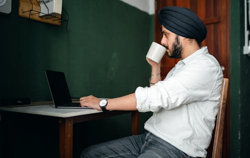 Side view of young bearded Indian male in casual wear and turban drinking coffee while working remotely on laptop