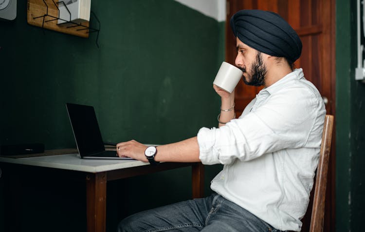 Focused Young Indian Man With Cup Of Coffee Working On Netbook At Home