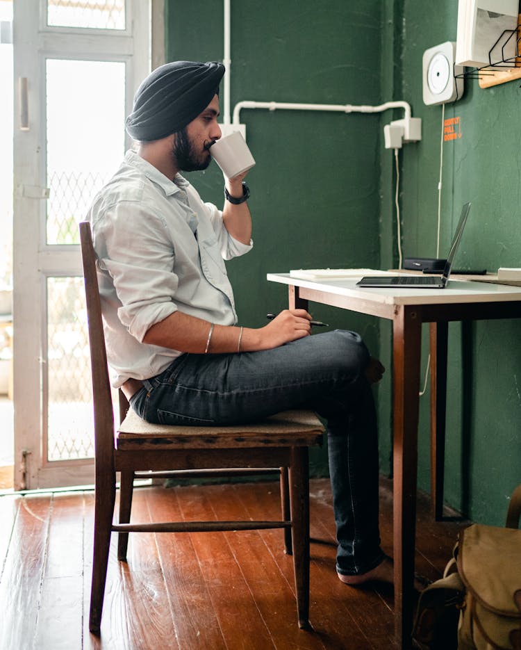 Young Indian Man Sitting At Table With Netbook And Drinking Coffee