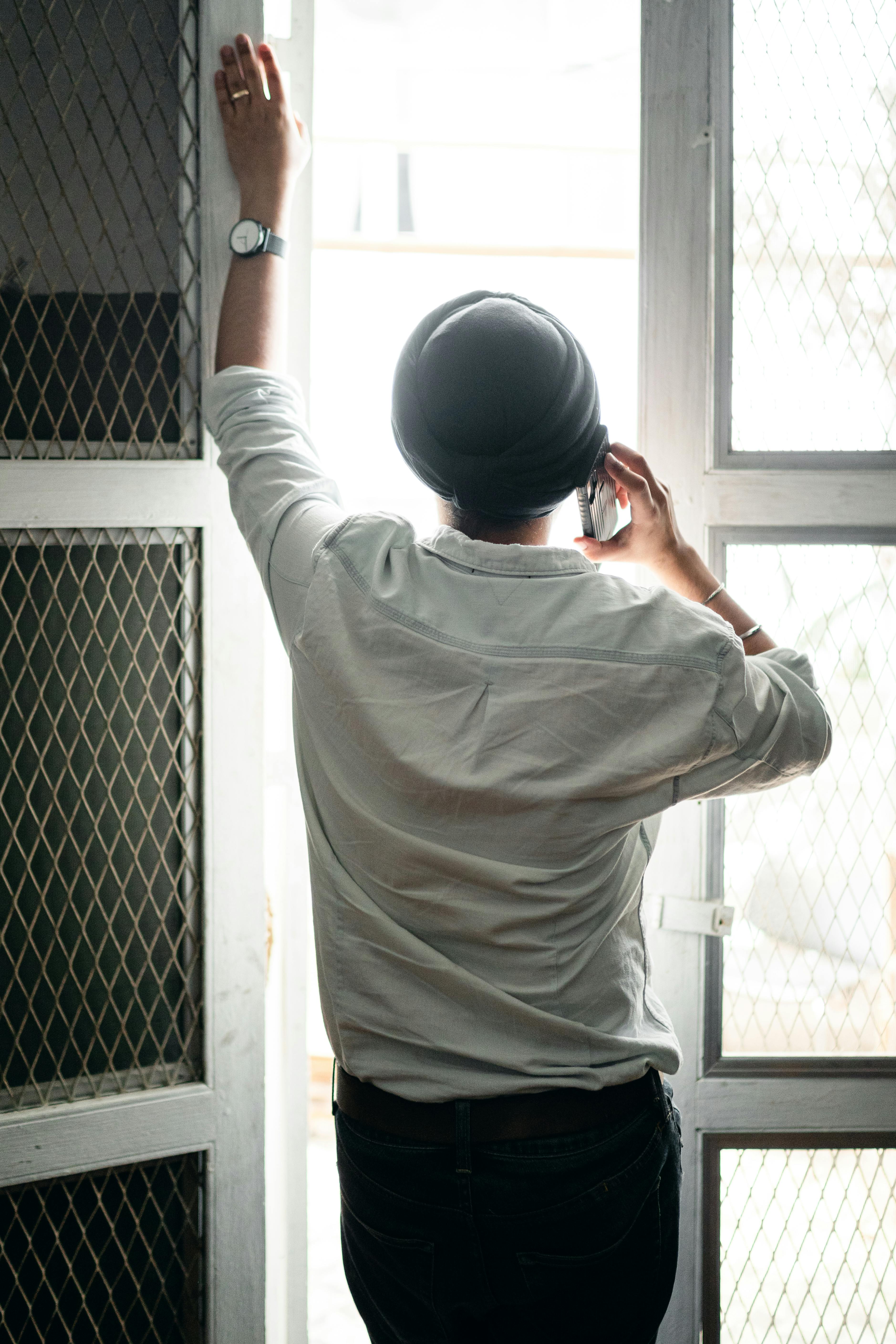 faceless young sikh man talking on smartphone standing near window