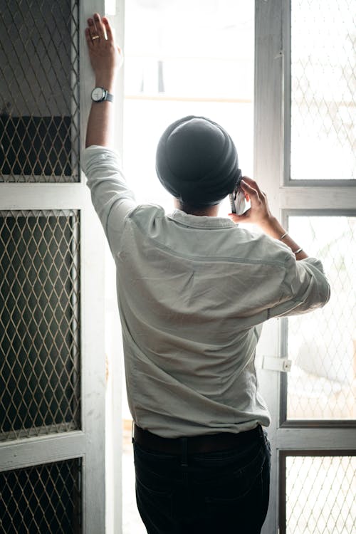 Back view of anonymous young Indian male in shirt and turban having phone conversation while standing against window