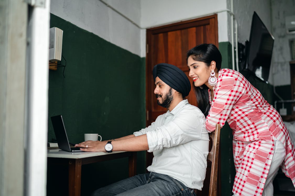 Free Side view of positive young Sikh man in shirt and turban working on laptop at home while wife leaning on chair behind Stock Photo