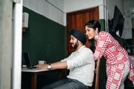 Side view of positive young Sikh man in shirt and turban working on laptop at home while wife leaning on chair behind