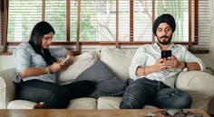 一个戴着头巾、留着严肃胡须的年轻男人和一个穿着休闲服装的女人，正坐在舒适的沙发上用智能手机发信息