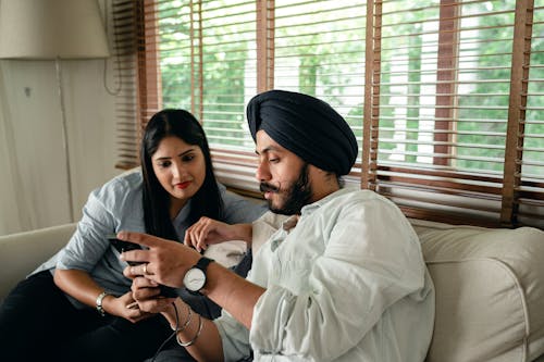 Focused young married Indian couple working on project using smartphone while sitting on comfortable couch at home