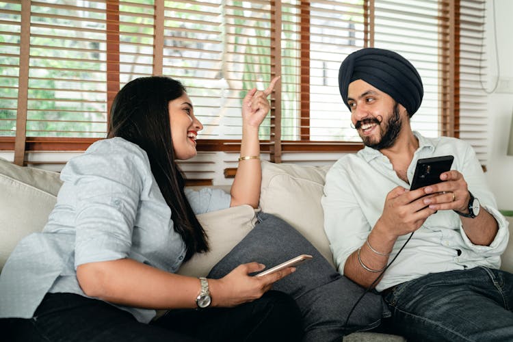 Happy Indian Couple With Mobile Phones Having Fun On Couch