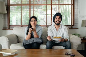 Positive male with plate of food and female in casual clothes sitting on sofa in living room and watching TV at home