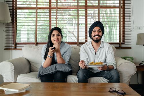 Free Positive male with plate of food and female in casual clothes sitting on sofa in living room and watching TV at home Stock Photo