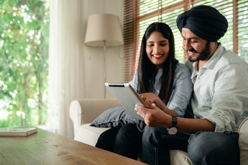 Optimistic ethnic male and female in casual outfit sitting on sofa in living room in daytime and using tablet together