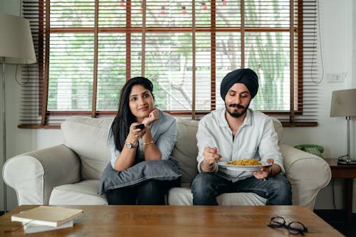 Pensive Indian male having meal on sofa while woman female in casual clothes sitting near and switching TV channels