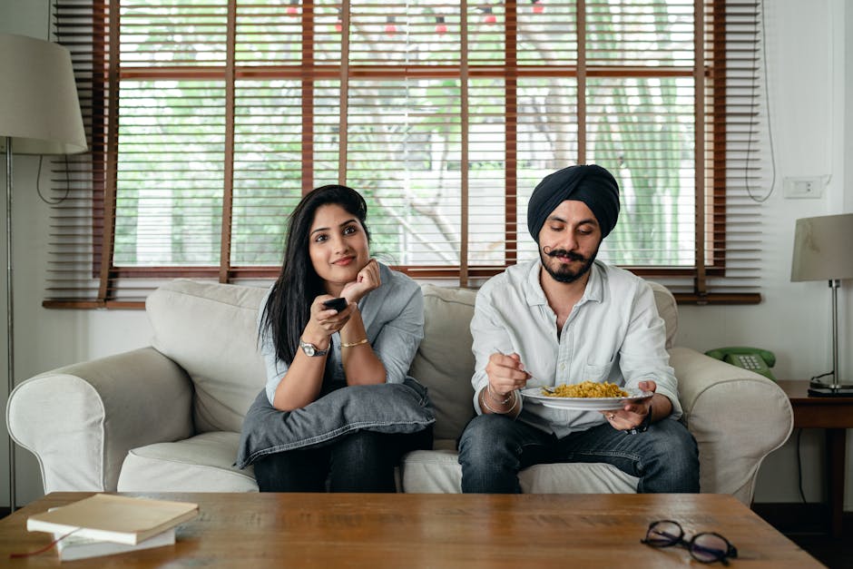 Pensive Indian male having meal on sofa while woman female in casual clothes sitting near and switching TV channels