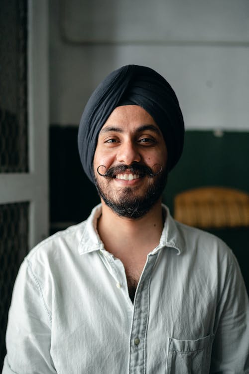Free Positive bearded Indian male in shirt and turban standing in room and smiling while looking at camera Stock Photo