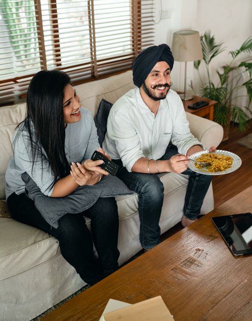 Free From above of content Indian wife with remote control and husband in turban with plate of food sitting on sofa in living room Stock Photo