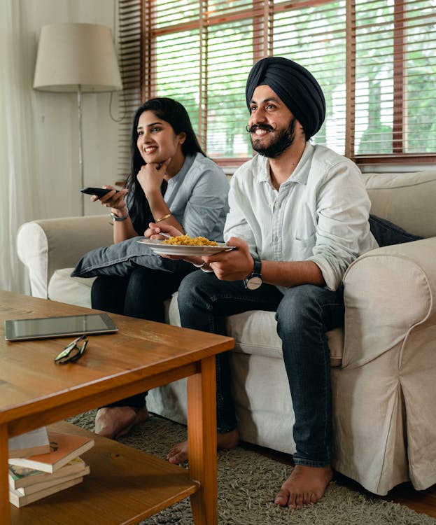 Free Happy Indian female and bearded male in casual clothes sitting on sofa in living room and smiling while watching tv Stock Photo