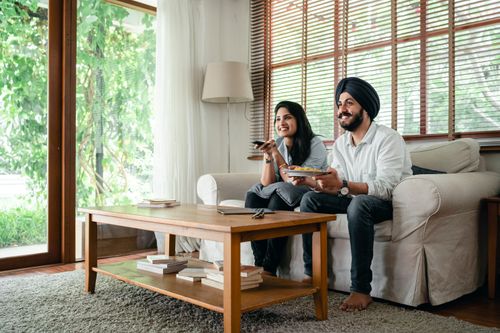 Free Cheerful Indian woman and man in turban spending free time together while watching television and sitting on sofa in living room near table Stock Photo