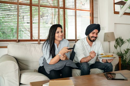 Free Happy ethnic male with plate of food and female with remote control sitting on couch in living room while watching movie Stock Photo