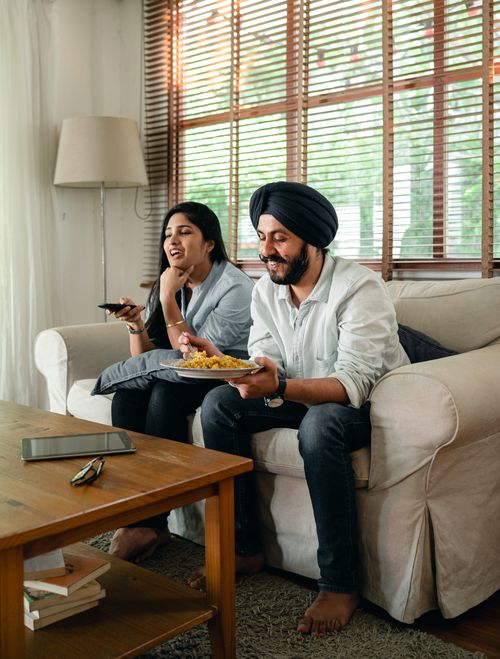 Free Happy Indian male having meal and female with remote control in hand watching tv and sitting on sofa in living room Stock Photo
