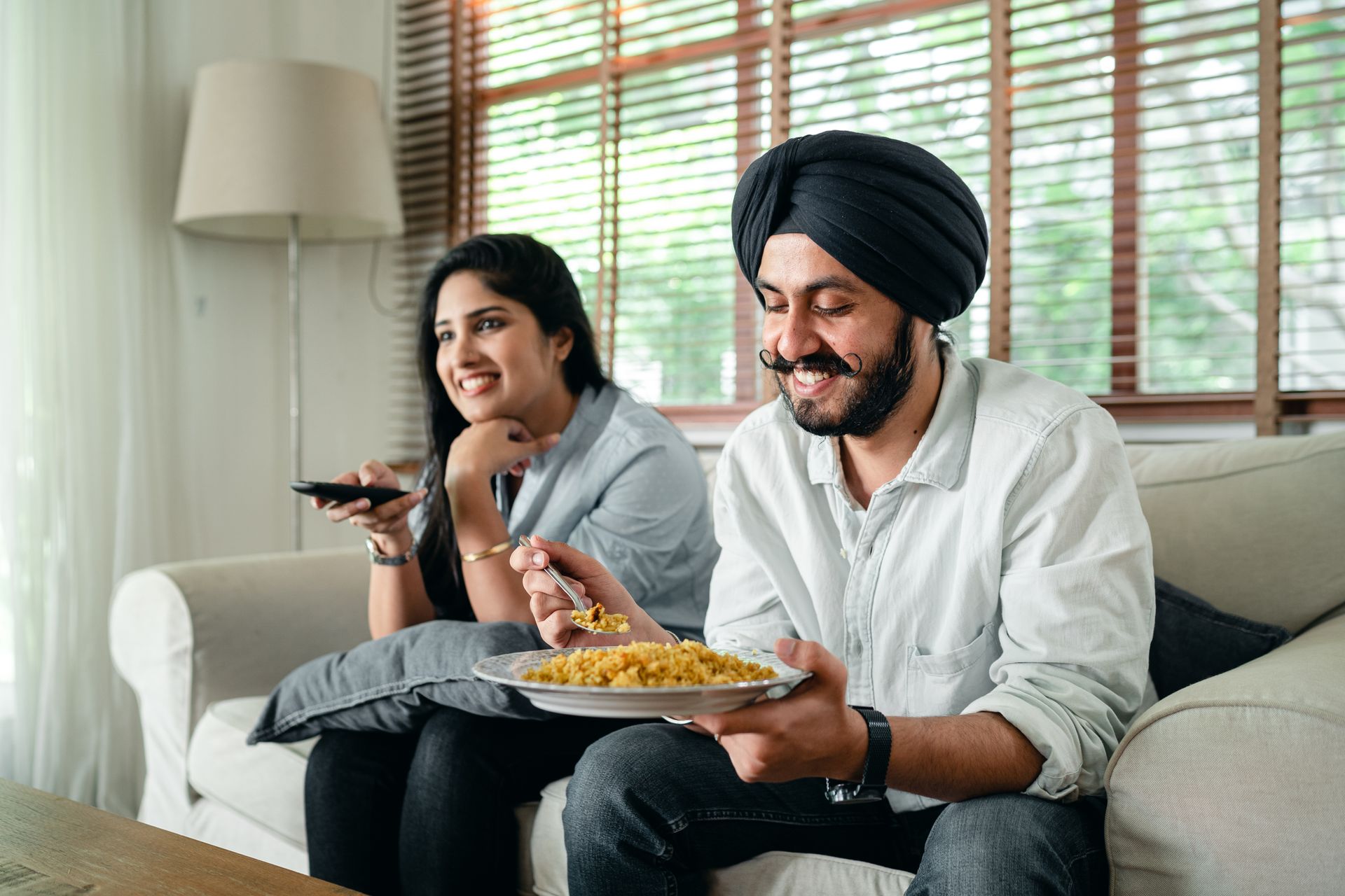 Optimistic Indian bearded male in turban and female with long hair sitting on sofa and spending leisure time together while watching television