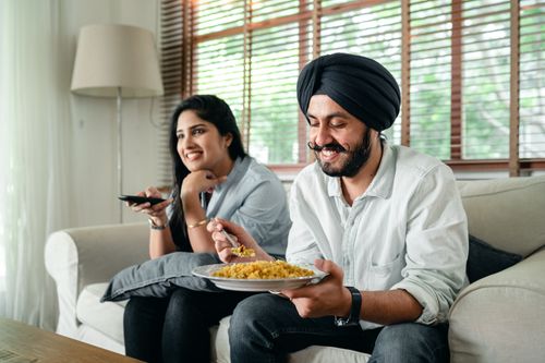 Free Optimistic Indian bearded male in turban and female with long hair sitting on sofa and spending leisure time together while watching television Stock Photo