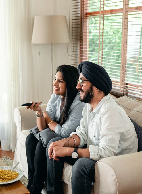 Free Side view of cheerful Indian male in eyeglasses sitting with female with remote control and sitting on couch in living room while watching movie Stock Photo