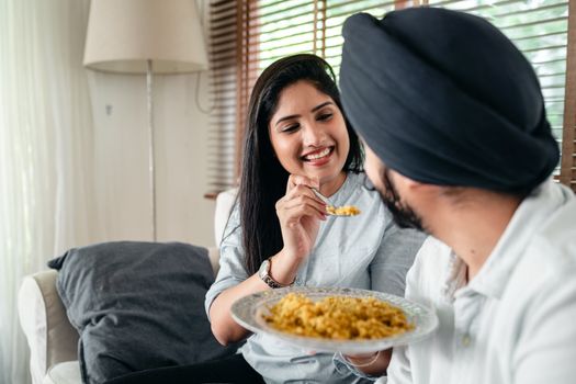 Happy Indian female in casual wear feeding beloved husband in blue turban while sitting on sofa in living room