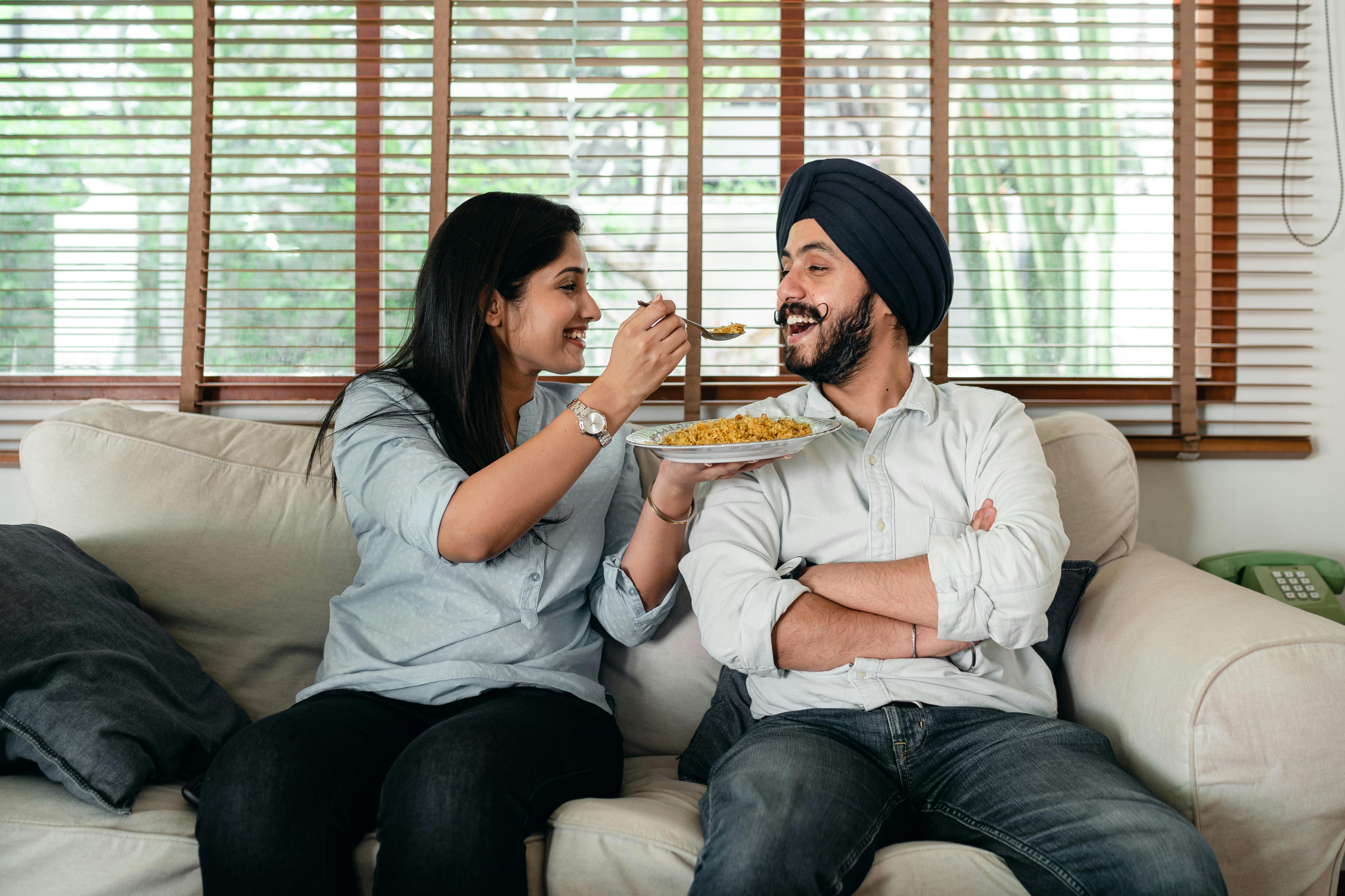 happy woman feeding ethnic man while sitting on sofa