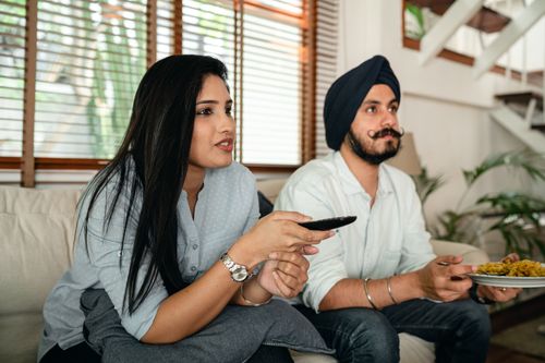 Free Calm Indian woman with remote control and man in turban and with mustache having meal while watching TV on couch Stock Photo