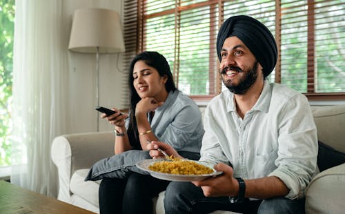 Positive young bearded man in casual wear and turban eating traditional saffron rice on sofa while wife switching channels on TV with remote controller