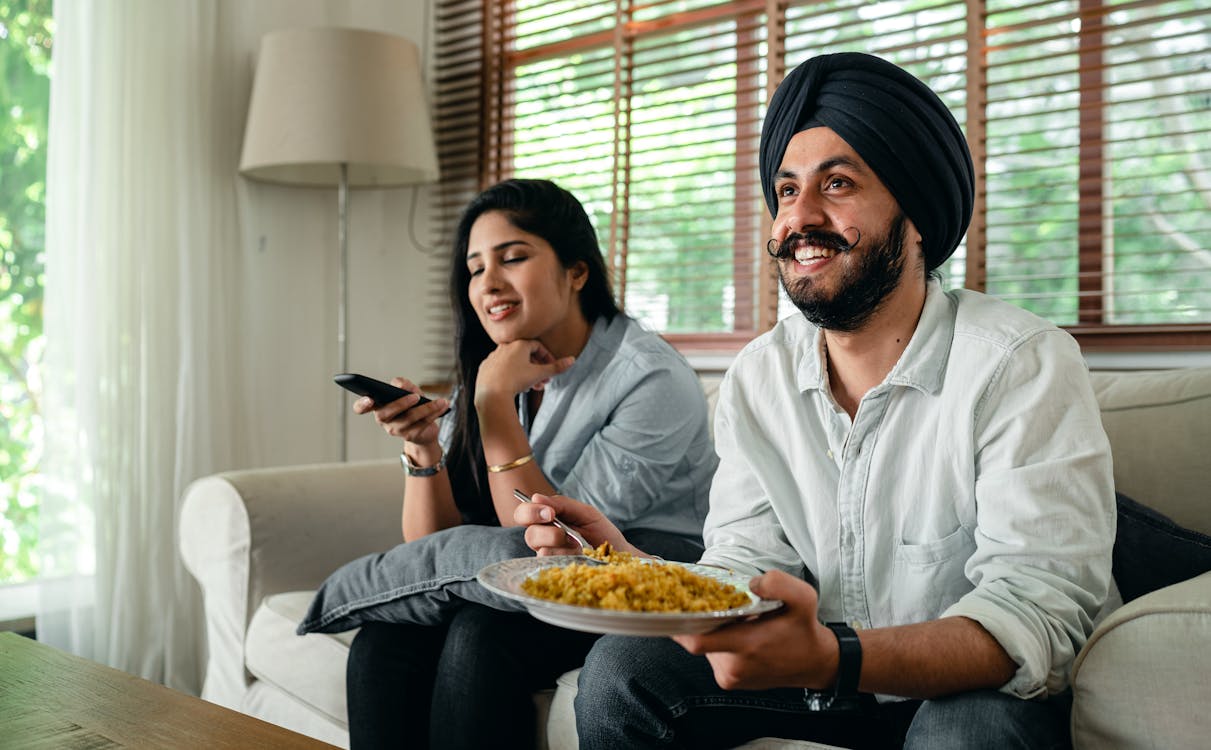 Free Positive young bearded man in casual wear and turban eating traditional saffron rice on sofa while wife switching channels on TV with remote controller Stock Photo