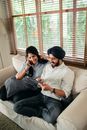 From above of cheerful young Sikh man in casual clothes and turban relaxing on sofa and browsing tablet while wife talking on smartphone