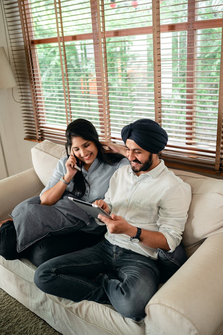 Young Indian Couple Using Tablet And Having Phone Conversation On Couch
