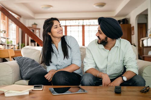 Cheerful young Indian man and woman in casual clothes smiling and looking at each other while sitting on sofa at home