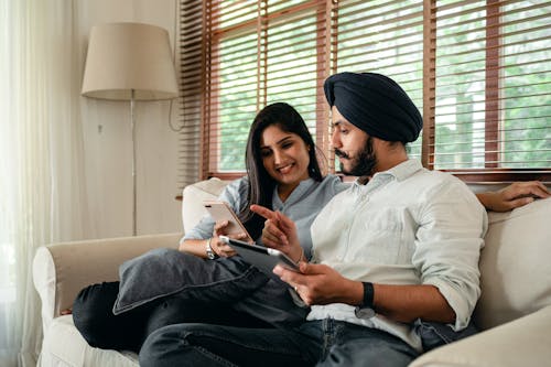 Young smiling Indian woman using smartphone and showing photo to husband working on tablet