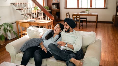 From above of joyful cuddling young Indian couple joking and laughing while resting together on comfortable sofa