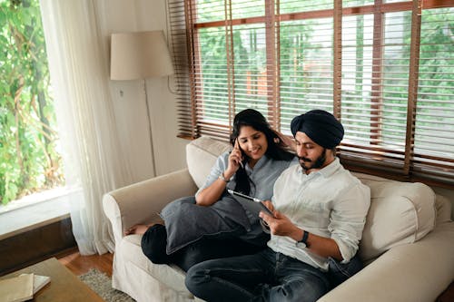 Young Indian man browsing tablet while woman talking on smartphone on couch