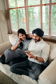 From above of young Indian man and woman in casual clothes sitting together on sofa near window in cozy apartment and browsing tablet while woman talking on mobile phone